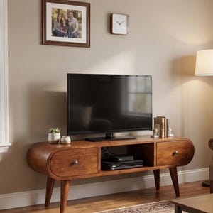 May include: A mid-century modern style TV stand in a warm brown finish. The stand features two drawers with round knobs, an open shelf for media components, and tapered legs. A large flat-screen TV sits on top, with a framed family photo and a square clock on the wall.