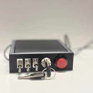 May include: A black control box with a key switch, three toggle switches, and a red button. The box has a rectangular shape and a dark gray frame. A key is inserted in the key switch. Wires extend from the box.