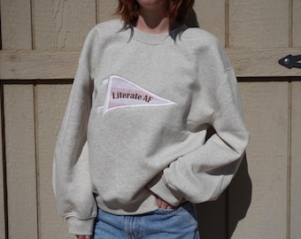 Könnte beinhalten: Ein beigefarbener, melierter Rundhals-Pullover mit einer weißen, wimpelförmigen Grafik auf der Brust. Der Wimpel enthält ein rosa Streifenmuster und den in Dunkelbraun gedruckten Text Literate AF. Der Pullover hat lange Ärmel.
