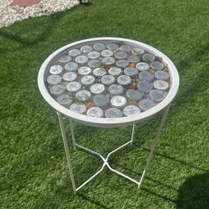以下が含まれることがあります： 白い金属フレームと、銀色のボトルキャップと小さな花が埋め込まれた透明な天板を備えた丸いサイドテーブル。 テーブルは十字型のベースで、緑の芝生の上に置かれています。