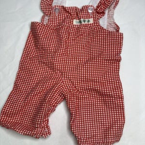 May include: Red and white checkered baby overalls with white ruffled straps and a small tag. The overalls have button closures and are made of a lightweight fabric. The garment is designed for infants and toddlers.
