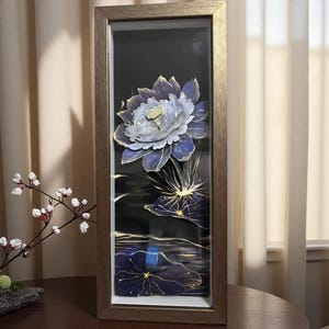 May include: A framed artwork featuring a detailed, layered paper lotus flower in shades of blue and white, with gold accents. The artwork is set against a black background, with reflections of the flower and lily pads. The frame is a warm, light brown.