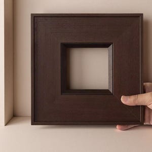 May include: A square, dark brown wooden frame with a smooth, matte finish and a bevelled inner edge. The frame features a clean, minimalist design with a wide border and a hollow centre, showcasing the natural grain texture of the dark wood material.