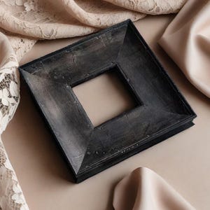 May include: A square, dark gray picture frame with a distressed finish. The frame has a square opening in the center and is set against a neutral background with draped fabric.