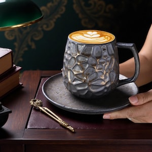 May include: A gray ceramic mug with a textured, stone-like pattern and gold accents, filled with coffee topped with latte art. The mug sits on a matching saucer, held by a person's hands. A decorative gold-colored pen is on the table.