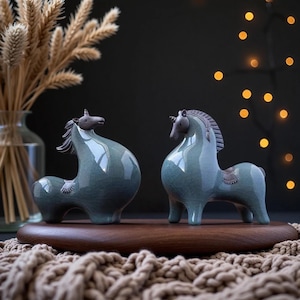 May include: Two ceramic unicorn figurines with a glossy, crackled blue-green glaze. Each figure features a dark brown head, mane, and tail, with a smooth, rounded body shape. The unicorns are positioned side-by-side on a circular wooden base.