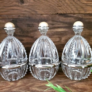 May include: Three clear glass egg-shaped containers with silver lids and a pearl-like knob on top. The containers are sitting on a wooden surface.