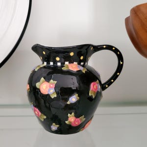 May include: Black ceramic pitcher with a floral pattern in pink, orange, and purple. The pitcher has a curved handle and a spout. The rim is decorated with polka dots and a checkered pattern. A decorative piece for the kitchen.