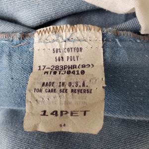 Op de afbeelding: Close-up van lichtblauwe denimstof met een aangehecht label. Het label luidt "50% COTTON, 50% POLY, MADE IN U.S.A." en bevat andere details. De stof is licht gekreukt.