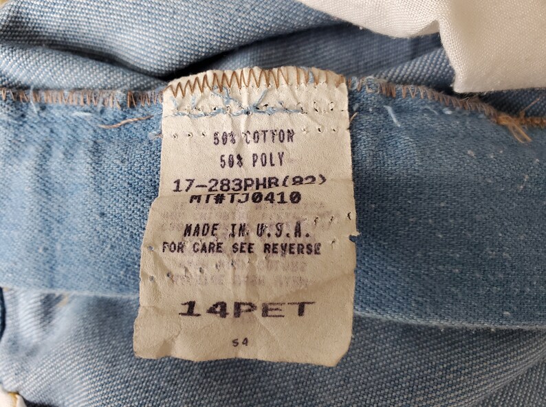 Op de afbeelding: Close-up van lichtblauwe denimstof met een aangehecht label. Het label luidt "50% COTTON, 50% POLY, MADE IN U.S.A." en bevat andere details. De stof is licht gekreukt.