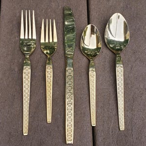 May include: A set of five gold-colored flatware pieces, including forks, a knife, and spoons. The handles feature a detailed floral pattern. The utensils are arranged on a dark brown surface, showcasing their elegant design.