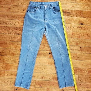 Op de afbeelding: Lichtblauwe jeans met een rechte pijp. De jeans heeft een hoge taille en een klassieke vijf-zakken stijl. Een gele meetlint ligt naast de jeans en toont de lengte in centimeters.