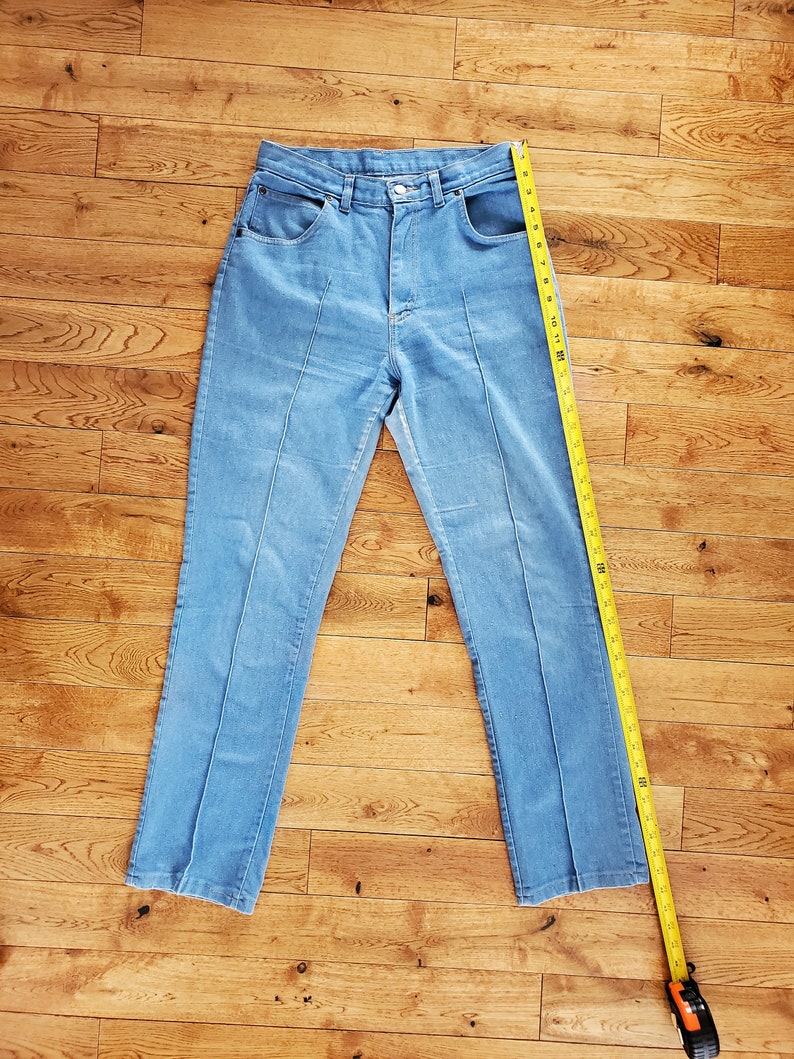 Op de afbeelding: Lichtblauwe jeans met een rechte pijp. De jeans heeft een hoge taille en een klassieke vijf-zakken stijl. Een gele meetlint ligt naast de jeans en toont de lengte in centimeters.