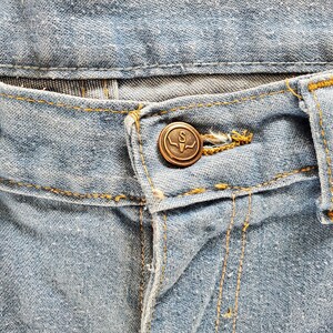 Op de afbeelding: Close-up van een lichtblauwe spijkerbroek. De jeans heeft een bronzen knoop met een gestileerde "S" en een stierenkopontwerp. Gele stiksels accentueren de naden en riemlussen.