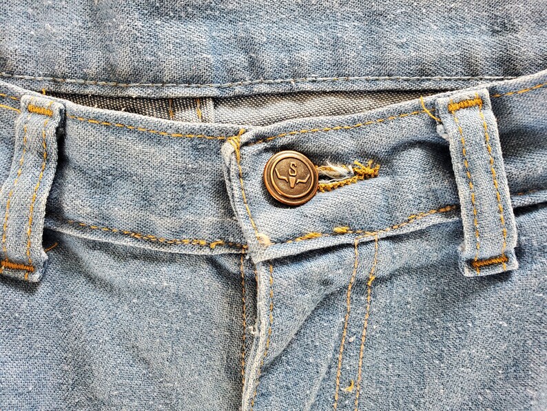 Op de afbeelding: Close-up van een lichtblauwe spijkerbroek. De jeans heeft een bronzen knoop met een gestileerde "S" en een stierenkopontwerp. Gele stiksels accentueren de naden en riemlussen.