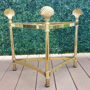 May include: A triangular gold-toned metal stand featuring three vertical legs, each topped with a decorative shell-shaped finial. The frame consists of two horizontal triangular tiers connected by the legs, which terminate in small, rounded feet.