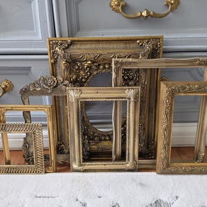 May include: A collection of six rectangular and oval picture frames in various shades of gold. The frames feature ornate, embossed, and textured detailing along their borders, ranging from intricate floral patterns to simple rope-like designs.