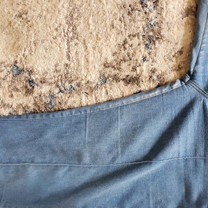 Op de afbeelding: Lichtblauwe denim stof, mogelijk van een spijkerbroek, met zichtbare stiksels en een getextureerd uiterlijk. De stof is gevouwen, met de naad en de richting van de snit van het kledingstuk. De achtergrond is een beige en bruin patroon tapijt.