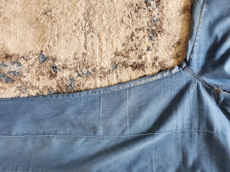 Op de afbeelding: Lichtblauwe denim stof, mogelijk van een spijkerbroek, met zichtbare stiksels en een getextureerd uiterlijk. De stof is gevouwen, met de naad en de richting van de snit van het kledingstuk. De achtergrond is een beige en bruin patroon tapijt.