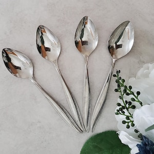 May include: Four silver teaspoons with rounded bowls and long, slender handles. The spoons are arranged in a fan shape on a white surface.
