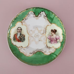 May include: A green and gold porcelain saucer with a white center featuring two portraits. The portraits are of a man in a military uniform and a woman in a pink dress.