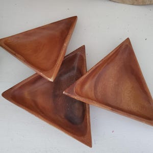 May include: Four wooden, triangular serving dishes. The dishes are a warm brown color with visible wood grain. The edges are slightly raised, creating a shallow bowl shape. The dishes are arranged on a white surface.
