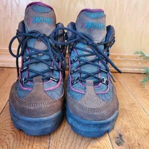 Peut inclure: Une paire de chaussures de randonn&eacute;e brunes et bleues avec une bordure bordeaux et le mot "BROOKS" sur le c&ocirc;t&eacute;.
