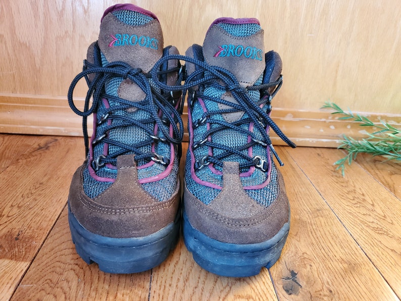 Peut inclure: Une paire de chaussures de randonn&eacute;e brunes et bleues avec une bordure bordeaux et le mot "BROOKS" sur le c&ocirc;t&eacute;.