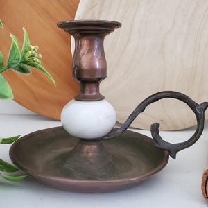 May include: A vintage-style candle holder with a copper-toned finish. It features a white marble sphere, a circular base, and a decorative handle. The candle holder is designed to hold a single candle.