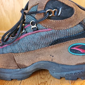 Peut inclure: Bottes de randonn&eacute;e en daim marron avec un logo turquoise et rouge qui dit "OUTDOOR". Les bottes ont un panneau en maille noire et une fermeture &agrave; lacets noire.