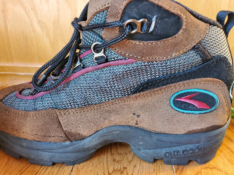 Peut inclure: Bottes de randonn&eacute;e en daim marron avec un logo turquoise et rouge qui dit "OUTDOOR". Les bottes ont un panneau en maille noire et une fermeture &agrave; lacets noire.