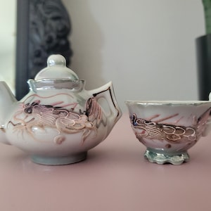 May include: A small, ornate porcelain tea set featuring a teapot and teacup. The set is primarily white with silver trim and detailed dragon designs in shades of pink, brown, and gold. The teacup has a small handle with gold accents.