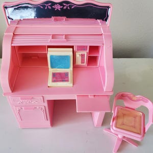 May include: A pink toy desk with a roll-top design, a computer, and a matching chair. The desk has a mirror on the top and a drawer on the bottom.