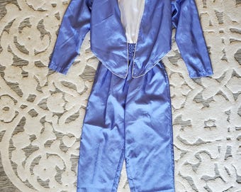 May include: A two-piece outfit consisting of a long-sleeved periwinkle blue jacket with a pointed hem and a matching pair of high-waisted trousers. The jacket is worn over a solid white sleeveless top. The fabric has a subtle sheen throughout.