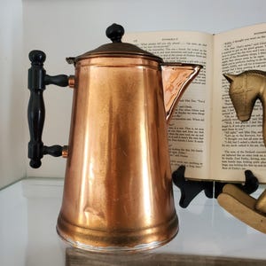 May include: A copper coffee pot with a black handle and lid. The pot has a shiny, reflective surface and a spout for pouring. The handle is made of wood and is attached to the side of the pot. The lid has a small, round knob on top.