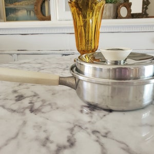 May include: A silver saucepan with a white handle and a matching lid. The saucepan is sitting on a white marble countertop.