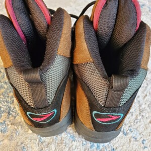 Peut inclure: Une paire de bottes de randonn&eacute;e en daim marron avec un maillage noir et un logo bleu et rose. Les bottes ont une languette de traction noire &agrave; l'arri&egrave;re de chaque botte.