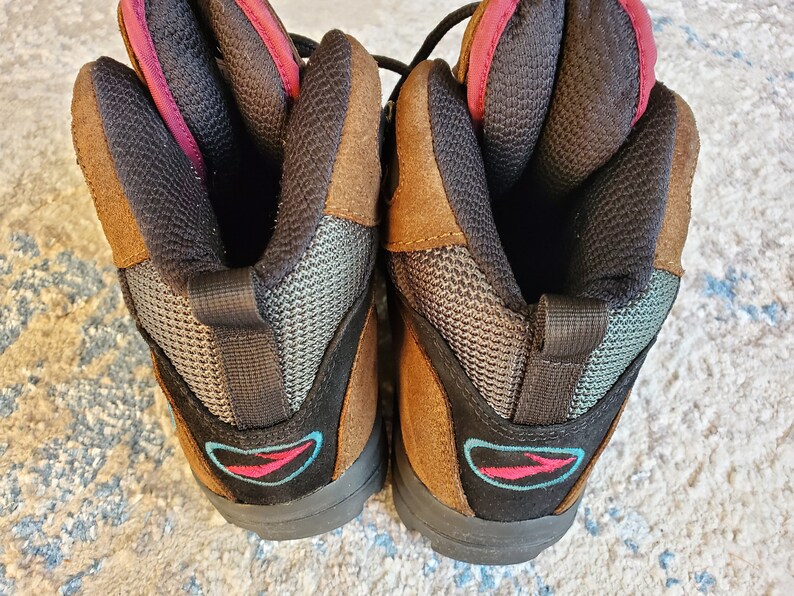 Peut inclure: Une paire de bottes de randonn&eacute;e en daim marron avec un maillage noir et un logo bleu et rose. Les bottes ont une languette de traction noire &agrave; l'arri&egrave;re de chaque botte.