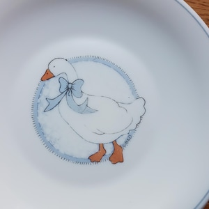 May include: A white ceramic plate with a blue and white illustration of a goose wearing a blue bow. The plate has a white background and a blue rim.