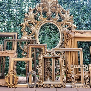 May include: A collection of various empty picture frames in gold and bronze tones, featuring ornate, carved, and smooth designs. The frames vary in size and shape, including rectangular and oval styles, arranged together in a layered display.
