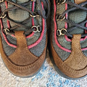 Peut inclure: Une paire de chaussures de randonn&eacute;e en daim marron avec des lacets noirs et des accents roses. Les chaussures ont une tige en mesh et une semelle en caoutchouc.
