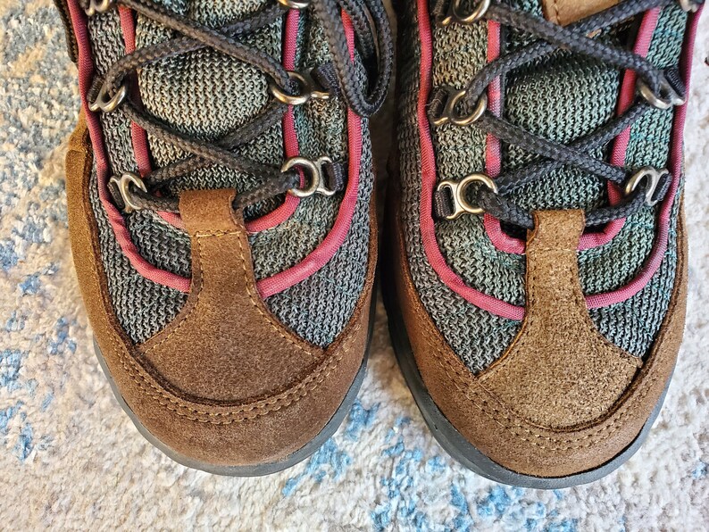 Peut inclure: Une paire de chaussures de randonn&eacute;e en daim marron avec des lacets noirs et des accents roses. Les chaussures ont une tige en mesh et une semelle en caoutchouc.