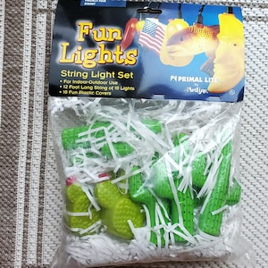 May include: A package of "Fun Lights" string light set. The package includes a 12-foot string of 10 lights with fun plastic covers. The lights are shaped like cacti. The packaging has the words "Fun Lights" in yellow and the brand name "Primal Lite".