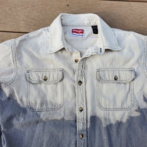 May include: A light blue denim shirt with a faded, ombre effect. The shirt has a button-up front, two chest pockets, and a Wrangler label on the front.