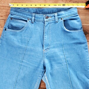 Op de afbeelding: Lichtblauwe denim jeans met een hoge taille en voorzakken. Een meetlint is over de bovenkant van de jeans geplaatst. De jeans heeft een knoopsluiting en een ritssluiting. De jeans is ongeveer 28 cm breed.