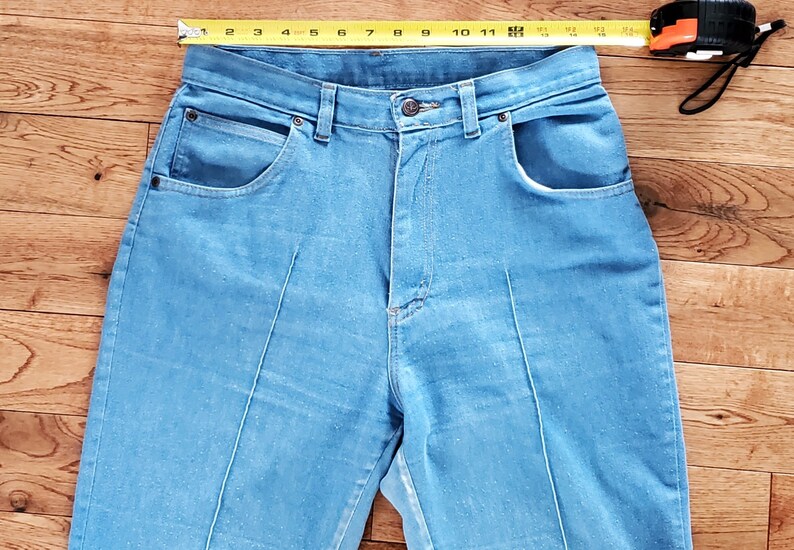 Op de afbeelding: Lichtblauwe denim jeans met een hoge taille en voorzakken. Een meetlint is over de bovenkant van de jeans geplaatst. De jeans heeft een knoopsluiting en een ritssluiting. De jeans is ongeveer 28 cm breed.