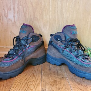 Peut inclure: Une paire de bottes de randonn&eacute;e brunes avec des lacets noirs et un panneau en mesh turquoise et bordeaux. Les bottes ont une semelle en caoutchouc noir et le mot "Brooks" est &eacute;crit sur le c&ocirc;t&eacute;.