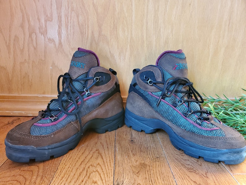 Peut inclure: Une paire de bottes de randonn&eacute;e brunes avec des lacets noirs et un panneau en mesh turquoise et bordeaux. Les bottes ont une semelle en caoutchouc noir et le mot "Brooks" est &eacute;crit sur le c&ocirc;t&eacute;.