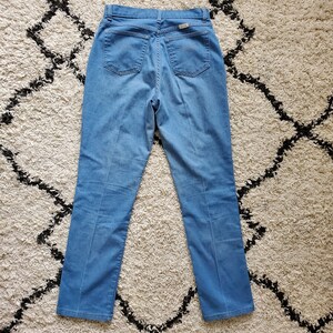 Op de afbeelding: Lichtblauwe spijkerbroek met hoge taille en rechte pijpen. De jeans heeft twee achterzakken en een klein label op de achterste tailleband. De jeans ligt plat op een patroonkleed.