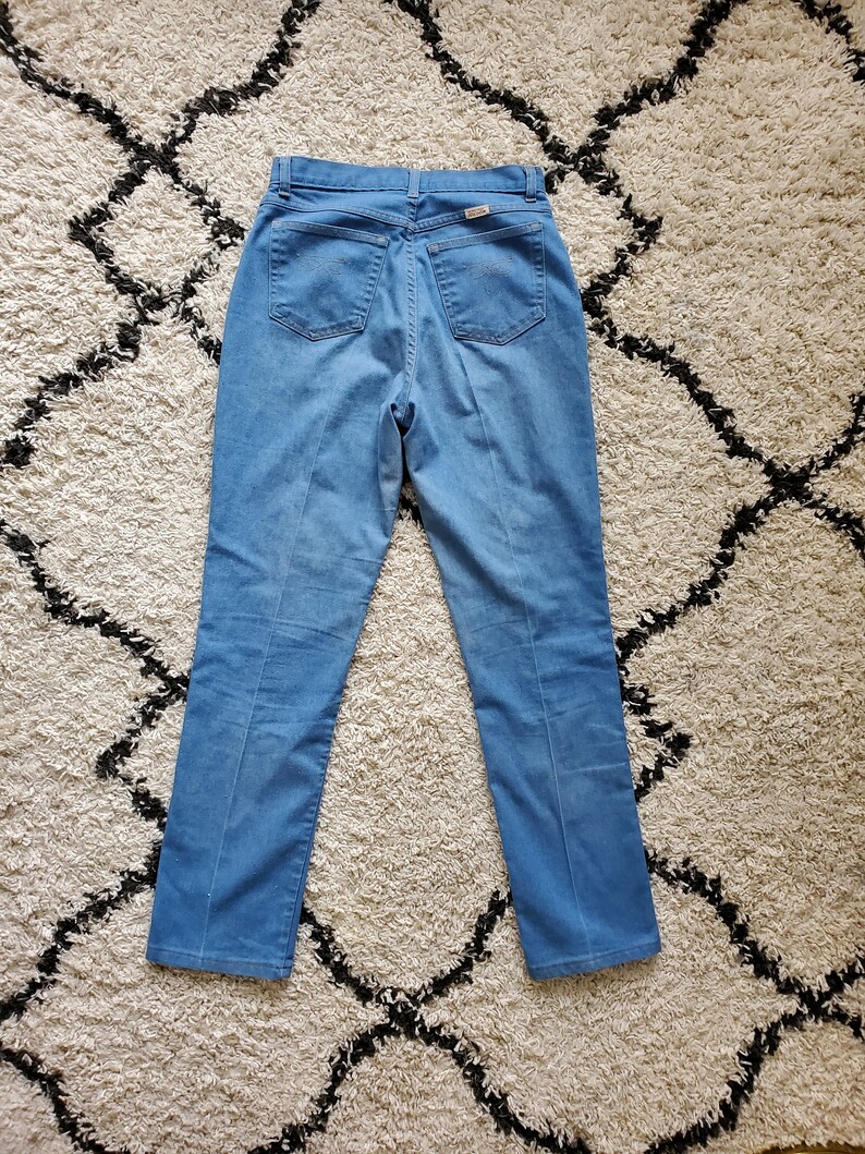 Op de afbeelding: Lichtblauwe spijkerbroek met hoge taille en rechte pijpen. De jeans heeft twee achterzakken en een klein label op de achterste tailleband. De jeans ligt plat op een patroonkleed.